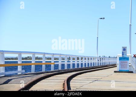 Eine Eisenbahn an der Küste mit Geländern und hölzerner Oberfläche und einem Warnschild vor einem klaren Himmel Stockfoto