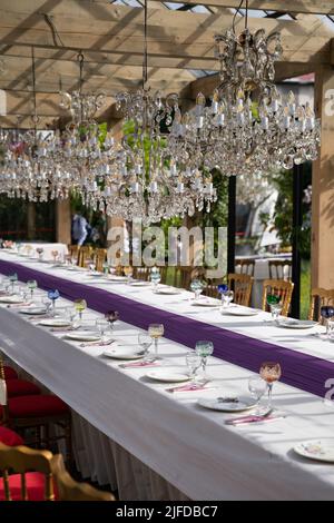 Wunderschöne Kristallleuchter mit Glühbirnen über dem gedeckten Festtisch mit Geschirr für die Zeremonie. Stockfoto