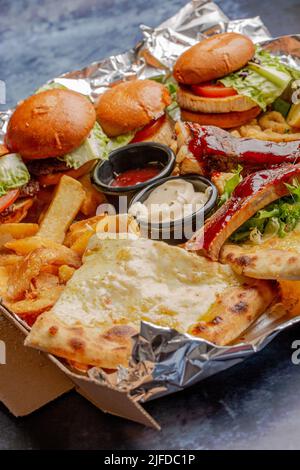 Eine Nahaufnahme von verschiedenen Fast Food-Sorten, darunter Burger, Pizza, Pommes auf Folie auf blauem Tisch Stockfoto