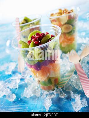 Plastikbecher gefüllt mit leckeren zum Mitnehmen sortierten reifen, gehackten Früchten, die mit Eis und Holzgabeln in einem hellen Raum auf den Tisch gelegt werden Stockfoto