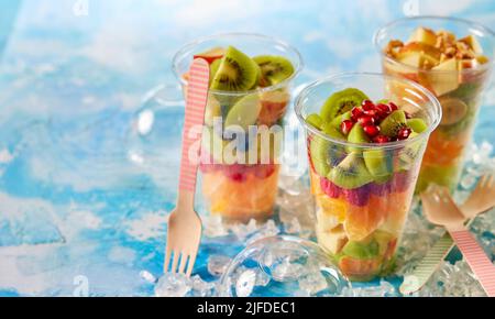 Mix aus frischen leckeren gehackten Früchten in Plastikbechern auf den Tisch mit Eis und Holzgabeln in der hellen Küche zu gehen Stockfoto