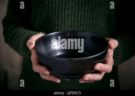 Crop anonyme Frau in warmen gestrickten grünen Pullover mit leeren schwarzen Keramikschale Stockfoto