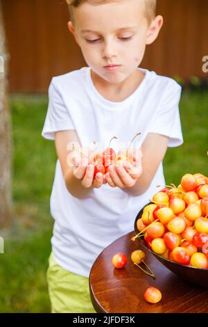 Ein sechsjähriger Junge steht auf einem grünen Rasen und spielt mit Kirschen. Sonniger Tag, grüner Rasen. Der blonde Junge schwelgt in Kirschen. Das Kind behandelt Kirschen. Stockfoto