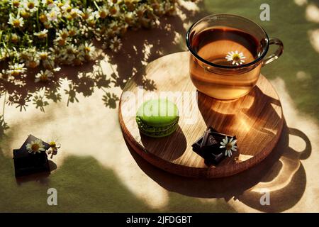 Ästhetische Teestunde im Freien, Kräutertee und Makarons-Dessert auf der Terrasse unter trendigen harten Schatten. Schöne Desserts, natürlicher Kamillentee - nachhaltiger ökologischer Lebensstil. Stockfoto