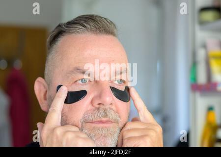 Mann mittleren Alters, der mit den Fingerspitzen Augengel-Feuchtigkeitspatches in einem Hautpflegekonzept anwendet Stockfoto