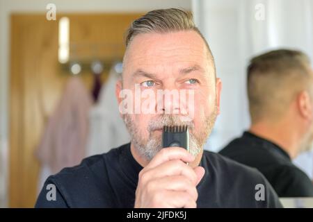 Nachdenklicher älterer Mann, der einen Trimmer an seinen Schnurrbart hält und mit einem nachdenklichen Gesichtsausdruck in Nahaufnahme zur Seite schaut Stockfoto