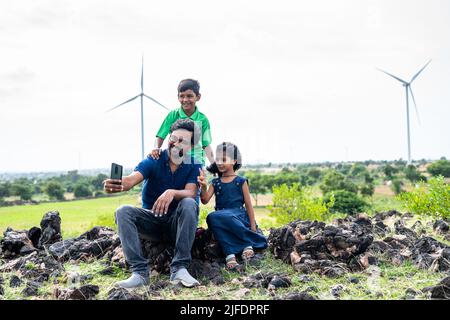 Glücklicher Vater und Kinder beim Selfie auf dem Handy vor Windturbinen oder Kraftwerk am Berg - Konzept des nachhaltigen Lebensstils, der Technologie Stockfoto