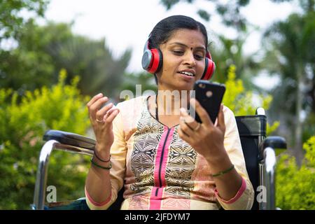 Frauen mit Behinderungen genießen es, auf kabellosen Kopfhörern Lieblingslieder zu hören, während sie im Park im Rollstuhl sitzen - Konzept der Entspannung, positiv Stockfoto