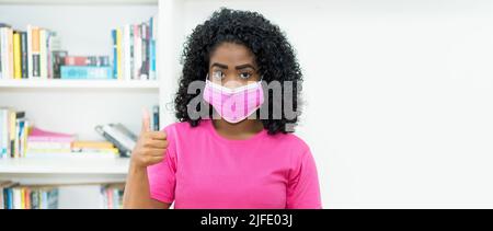 Optimistische afroamerikanische Frau mit rosa Gesichtsmaske als Schutz vor dem Covid-19-Virus Stockfoto