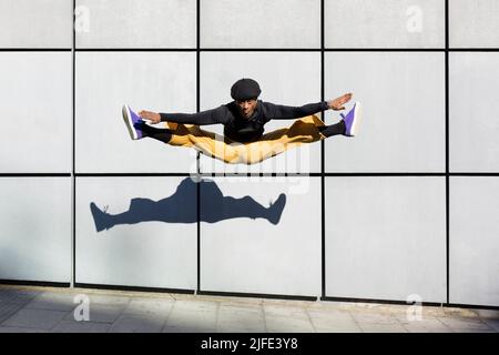 Afroamerikanischer junger Erwachsener springt energisch auf der Straße. Tänzerin, die Akrobatik in der Stadt macht. Moderner Look, Ästhetik urbaner Lifestyle. Sp Stockfoto