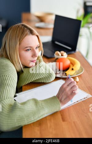 Die junge Frau denkt über etwas nach und macht sich zu Hause in der Küche Notizen. Stockfoto
