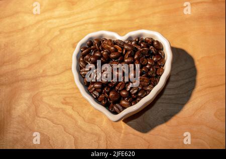 Eine Aufnahme von Kaffeebohnen über dem Kopf in einer herzförmigen weißen Schale auf einem strukturierten Holztisch - Stock-Fotografie Stockfoto