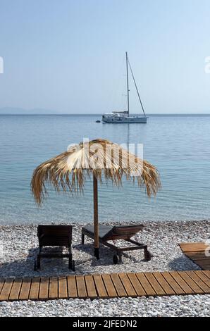 Strandszene, Strohschirm und Liegestühle und Jacht in der Ferne. Dorf Livadia, Insel Tilos, Dodcanese, Griechenland Stockfoto