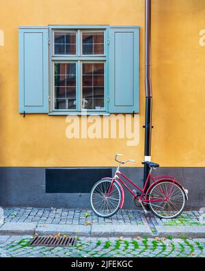 Rotes Fahrrad, das unter einem alten traditionellen Vintage-Fenster mit blauen Fensterläden in einer orangefarbenen Wand in einer engen Kopfsteinpflasterstraße in der Altstadt von Gamla Stan, Stockholm, Schweden, geparkt ist Stockfoto