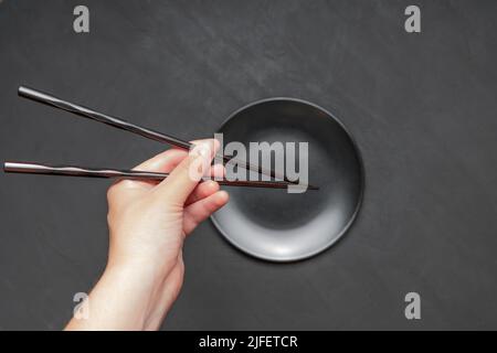 Ein Paar lackierte Essstäbchen in der Hand über einer leeren mattschwarzen Platte auf dunklem Hintergrund. Stockfoto