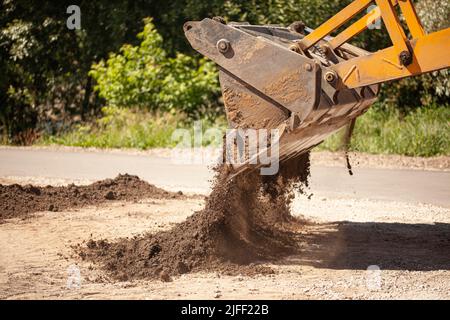 Der Baggereimer schüttet den Boden vor dem Hintergrund einer ländlichen Industriezone aus. Planierraupen auf der Baustelle. Stockfoto