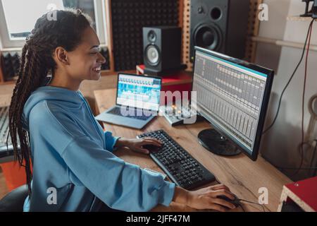 Schöne junge stilvolle afroamerikanische Toningenieurin und Produzentin in lässiger Kleidung, die im Musikstudio arbeitet und coole Songs kreiert Stockfoto