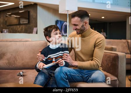 Ein hübscher, liebevoller junger Vater umarmte seinen Sohn sanft und spielte mit Spielzeugflugzeugen, während er auf die Zoll- und Passagierkontrolle wartete, bevor er den Flug besteigen konnte Stockfoto