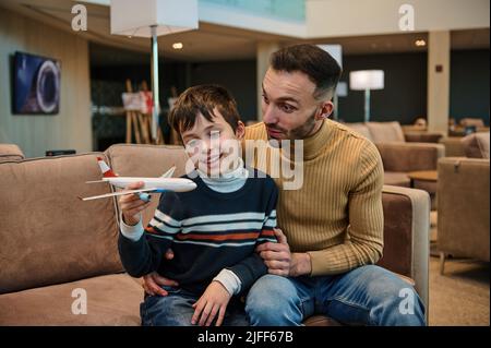 Schöner junger, liebevoller Vater, der mit seinem entzückenden Sohn ein Spielzeugflugzeug spielt, während er sich in der VIP-Lounge am Abflugterminal des internationalen Flughafens entspannt und wartet Stockfoto