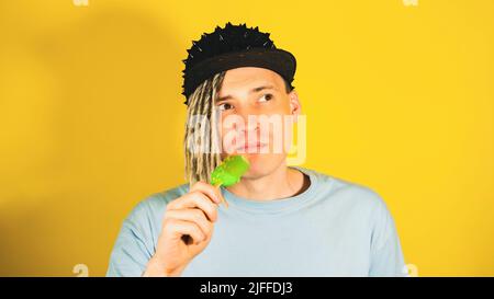 Junger, hübscher Mann mit Dreadlocks in schwarzer Mütze, der Eis auf gelbem Hintergrund isst. Verträumter Kerl, der Eisbecher isst und wegschaut Stockfoto