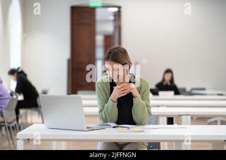 Glückliche, reife Studentin, die an einem virtuellen Kurs teilnimmt und sich das Webinar auf einem Laptop in der Bibliothek ansieht Stockfoto