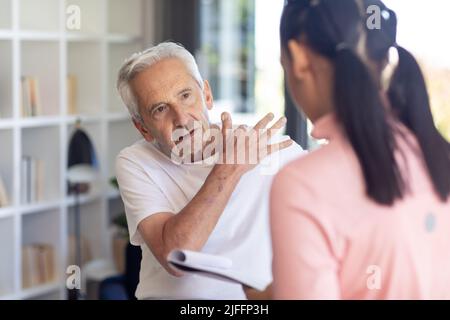 Kaukasischer älterer Mann, der der birazialen Physiotherapeutin sein Schulterschmerz-Problem zeigt Stockfoto