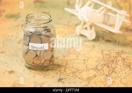 Glas mit Münzen und Reiseinschrift auf der Weltkarte mit Holzspielzeugflugzeug auf dem Tisch. Reisekonzept und Geld sparen. Stockfoto