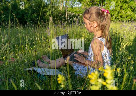 Kleines Mädchen, das auf Gras sitzt und einen Laptop benutzt. Bildung, Lifestyle, Technologiekonzept, Outdoor-Lernkonzept Stockfoto