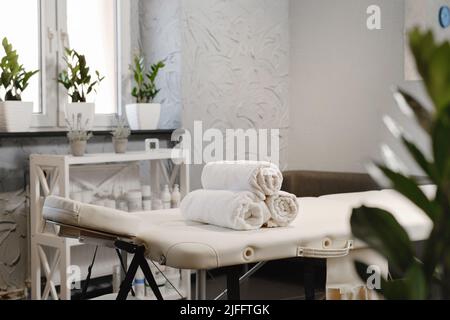 Im leeren Salon wurden weiße Handtücher auf den Massageliege gelegt. Das Innere der ästhetischen Klinik, niemand. Modernes Kabinett des Physiotherapeuten. Leerer Raum mit Pflanzen Stockfoto