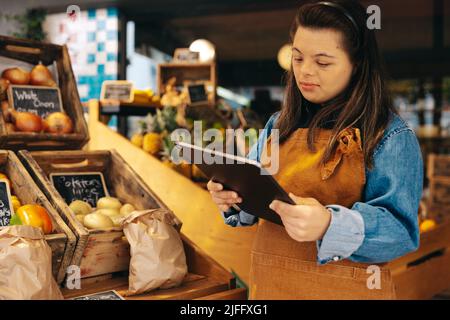 Ladenbesitzer mit Down-Syndrom mit einem digitalen Tablet im Frischwarenbereich eines Lebensmittelladens. Frau mit einer intellektuellen Behinderung, die ich arbeite Stockfoto