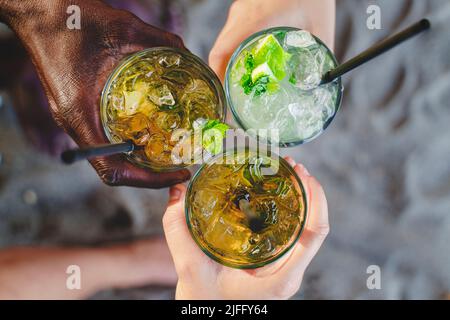 Nahaufnahme von multiethnischen Menschen, die einen frischen Mojito-Cocktail am Strand mit Sand im Hintergrund halten - Draufsicht - Ernteaufnahme - Menschen, Alkohol, vaca Stockfoto