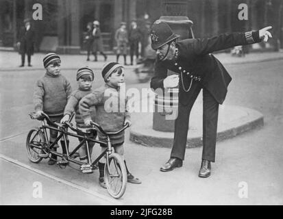 London im Jahr 1920s. Ein Polizist in einer Londoner Straße, der den drei Kindern auf einem Fahrrad den Weg erklärt. Das Fahrrad ist speziell für drei Personen gefertigt. Die Jungen sind in Wollpullover und Mützen gekleidet. Stockfoto