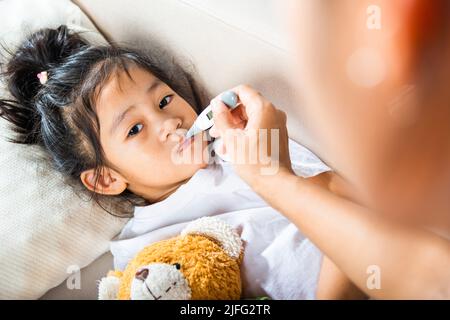 Krankes Kind. Mutter Eltern überprüfen die Temperatur ihrer kranken Tochter mit einem Digitalthermometer im Mund, Kind liegt im Bett und misst ihre Temperatur Stockfoto