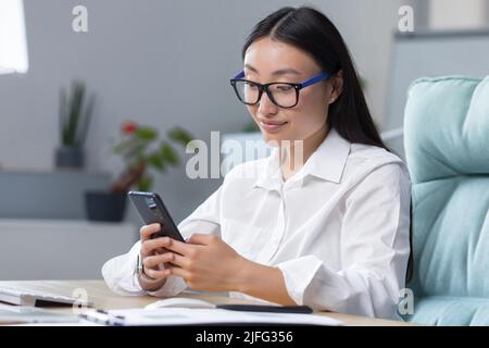Junge schöne asiatische Geschäftsfrau arbeitet im modernen Büro, nutzt Telefon, lächelt, liest gute Nachrichten, chattet in der App mit Freunden Stockfoto