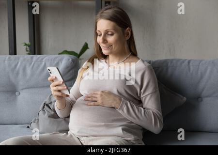 Die Schwangere benutzt das Mobiltelefon, das auf dem Sofa im Wohnzimmer sitzt Stockfoto