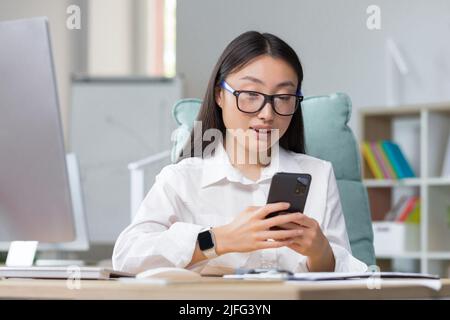 Junge schöne asiatische Geschäftsfrau arbeitet im modernen Büro, nutzt Telefon, lächelt, liest gute Nachrichten, chattet in der App mit Freunden Stockfoto