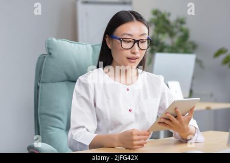Junge schöne asiatische Ärztin, die in einer modernen Klinik arbeitet, mit Tablet für Videoanrufe und Online-Konsultation von Patienten. Stockfoto