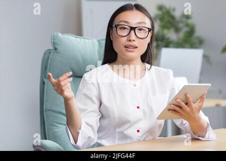Junge schöne asiatische Ärztin, die in einer modernen Klinik arbeitet, mit Tablet für Videoanrufe und Online-Konsultation von Patienten. Stockfoto