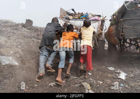 Drei schwarze Straßenjungen schoben einen schweren Müllwagen auf einer städtischen Deponie auf einen Haufen; informelle Abfallwirtschaft in Afrika Stockfoto