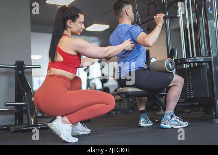 Junge kaukasische Personal Trainer Frau hilft Client auf Gym Station Anpassung zurück Stockfoto