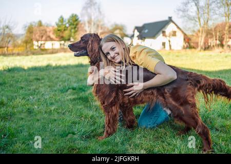 Happy 30s Frau lächelt, wenn sie im Sommer im Park mit ihrem roten irischen Setter-Hund auf frischem grünen Gras spielt Stockfoto