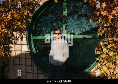 Stilvoller Mann in Sonnenbrille steht vor dem runden Fenster. Herbstwetter, gelbe und rote Blätter Stockfoto