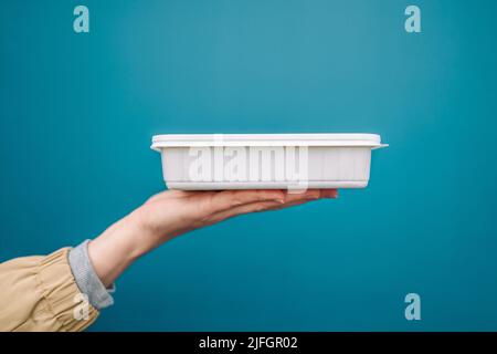 Frau Hände halten leere weiße Kunststoff Einweg-Lebensmittelbehälter Modell mit Kopie Platz auf blauem Hintergrund. Lieferungslebensmittelkonzept. Stockfoto