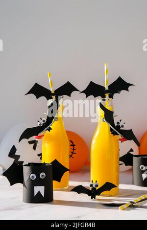 Halloween-Mocktails. Oranger alkoholfreier Cocktail für Halloween-Party auf weißem Tisch. Die Trinkhalme sind mit geschnittenen Papierfledermäusen verziert. Halloweens gruseliges dri Stockfoto