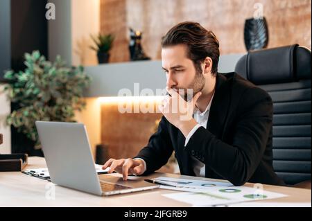 Fokussierter, erfolgreicher kaukasischer, intelligenter Geschäftsmann, Manager, Geschäftsleiter, sitzt am Arbeitsplatz im Büro, schaut nachdenklich auf den Laptop-Bildschirm, arbeitet an einem Geschäftsplan für ein neues Projekt Stockfoto