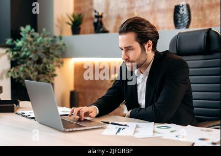 Ein kluger, seriöser kaukasischer Geschäftsmann, ein Finanzdirektor, sitzt an einem Schreibtisch in einem modernen Büro, konzentriert auf einem Laptop an einem neuen Projekt, entwickelt einen Geschäftsplan, eine Strategie, analysiert Risiken Stockfoto