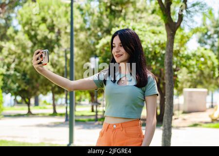 Brunette Millennial Frau trägt türkisfarbene T-Shirt im Stadtpark, im Freien ein Selbstporträt mit Smartphone. Sie schaut auf den Bildschirm und nimmt Sel Stockfoto