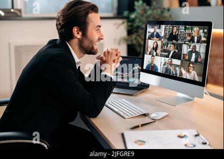 Erfolgreicher, positiver kaukasischer Geschäftsmann in einem Anzug, sitzt an einem Arbeitsplatz im Büro, verhandelt mit Partnern auf einer Videokonferenz mit einem Computer, diskutiert neues Projekt, Bedingungen der Zusammenarbeit Stockfoto