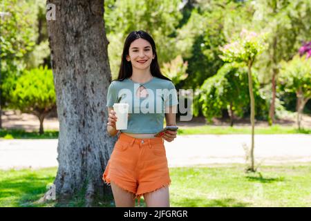 Junge Brünette Frau trägt türkisfarbenes T-Shirt und orangene Short im Stadtpark, hält im Freien Kaffeebecher zum Mitnehmen und nutzt Handy, während sie schaut Stockfoto