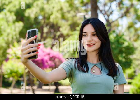 Glückliche Brünette Frau trägt türkisfarbenes T-Shirt im Stadtpark, im Freien ein Selbstporträt mit Smartphone. Sie schaut auf den Bildschirm und macht Selfie. Stockfoto